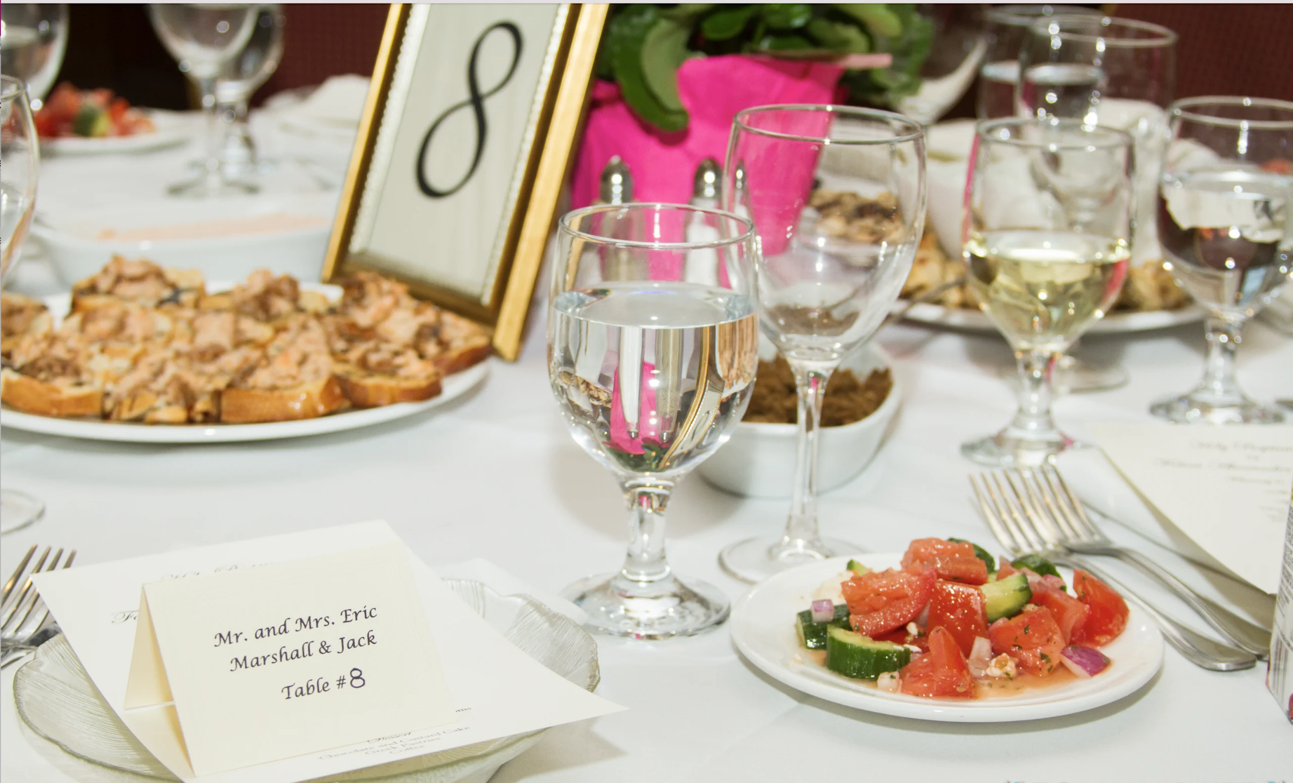 A table setting with salads, bruschetta, and drinks. A table 8 card, framed number, and menu are visible.