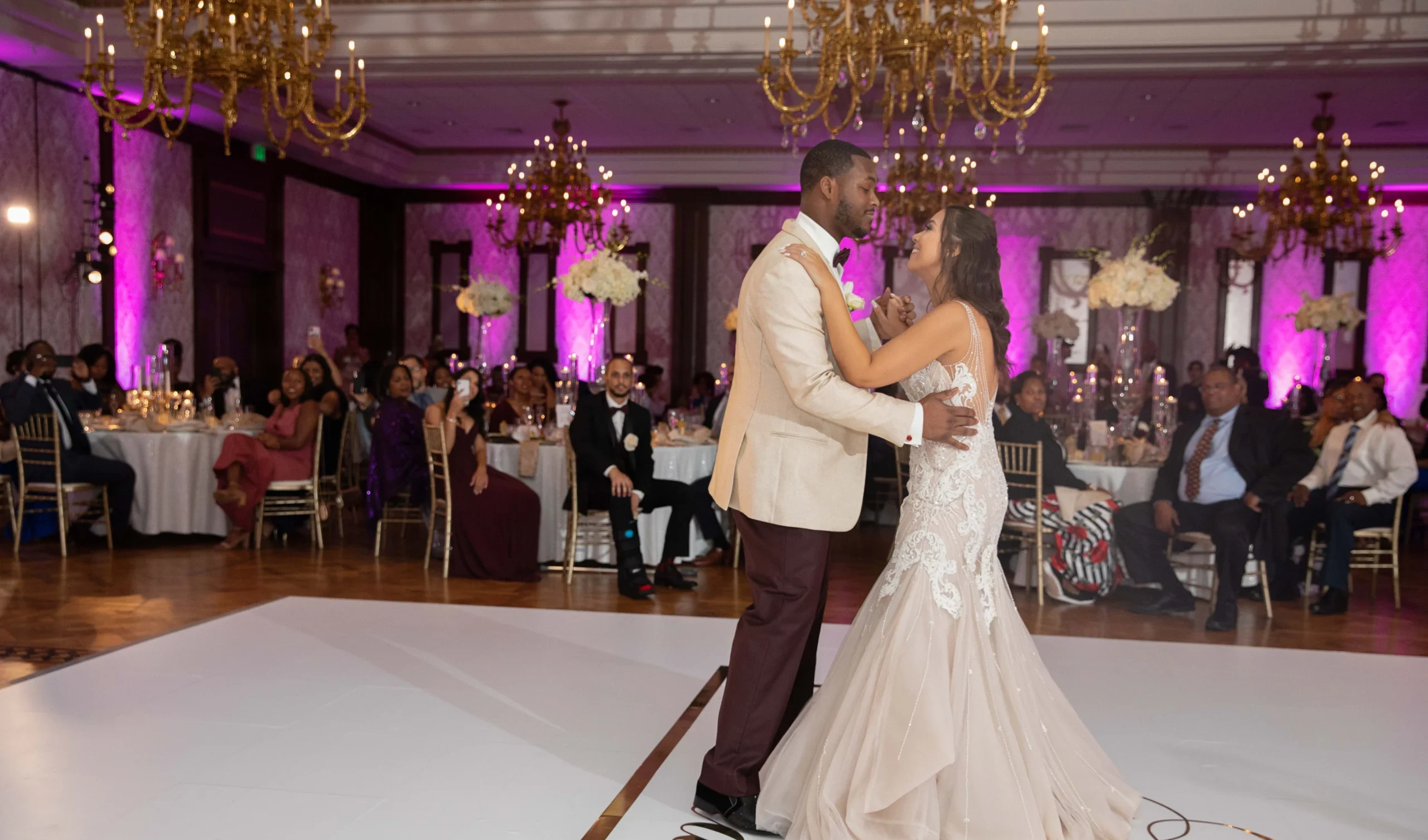 A couple dances together in an elegant ballroom with chandeliers and pink lighting, while guests watch from tables adorned with white flowers.