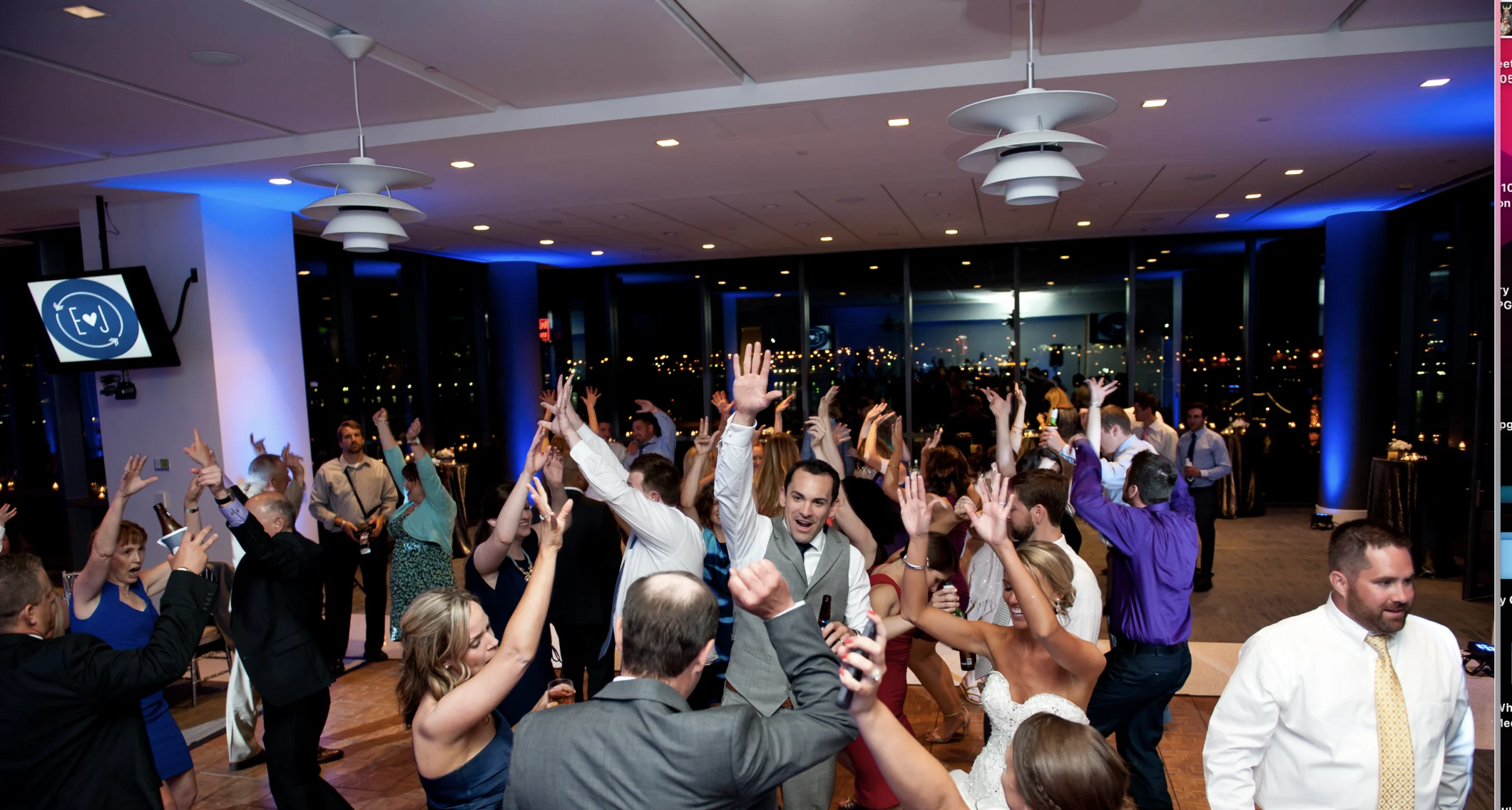 People are dancing with raised hands at a lively indoor event with city lights visible through large windows.