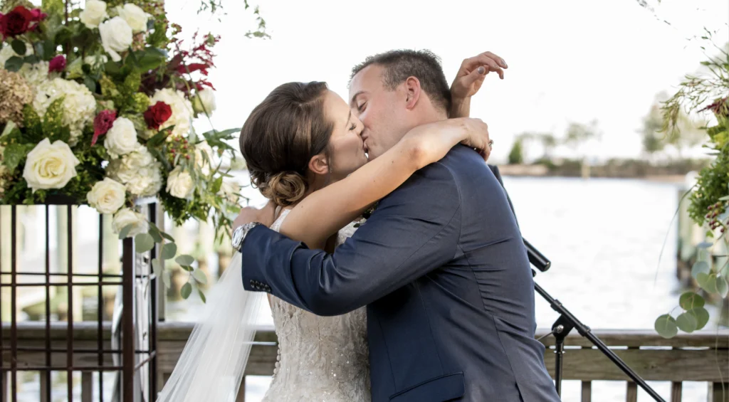 A bride and groom kiss at an outdoor wedding ceremony, surrounded by floral arrangements.