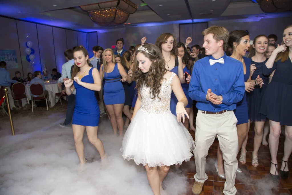 A group of young people in semi-formal attire dance in a foggy room with blue lighting. A girl in a white dress is in the center, surrounded by others wearing blue outfits.