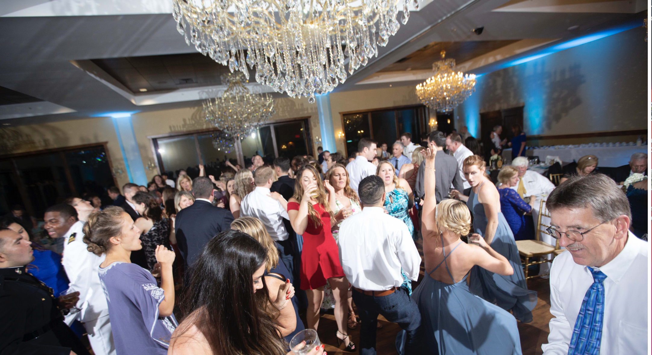 A large group of people dancing and socializing at a formal indoor event, with chandeliers overhead and a blue-lit ambiance.