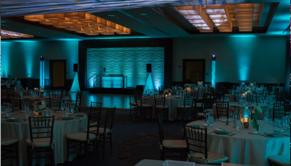 A dimly lit banquet hall with round tables set for an event. Blue uplighting decorates the walls and a DJ booth is at the center. Ceiling features patterned panels.