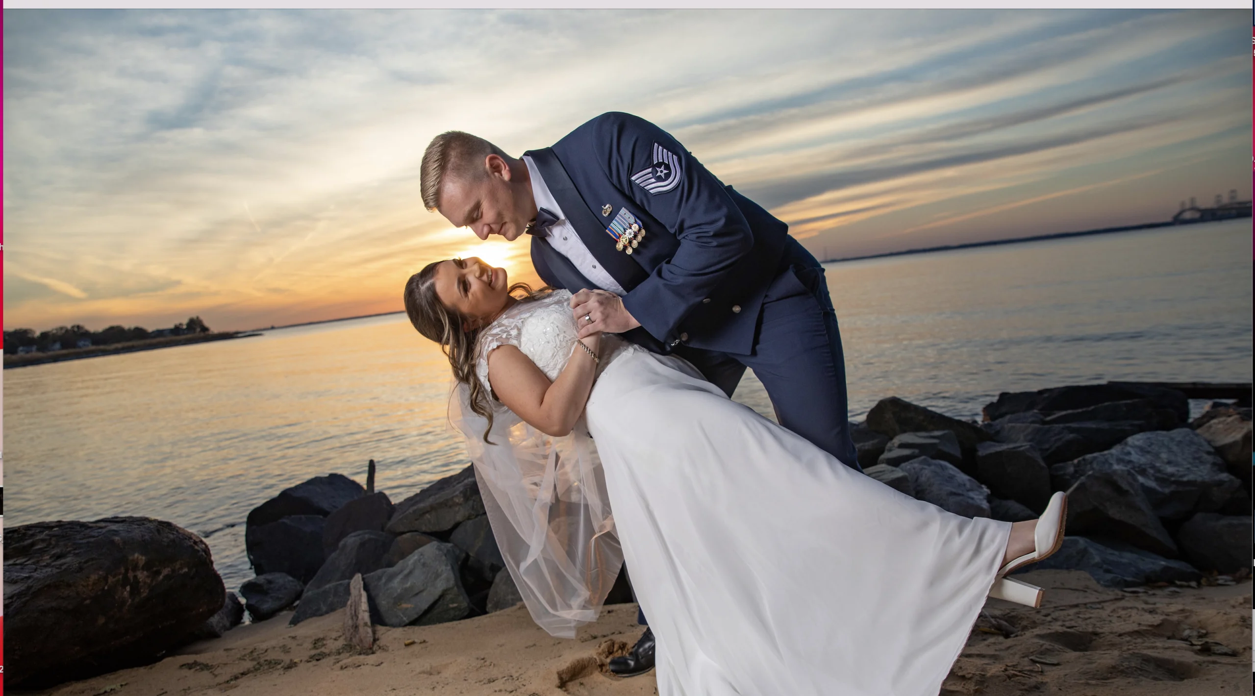 A couple poses on a beach at sunset; the man in a military uniform dips the woman, who is wearing a white wedding dress, near the water.