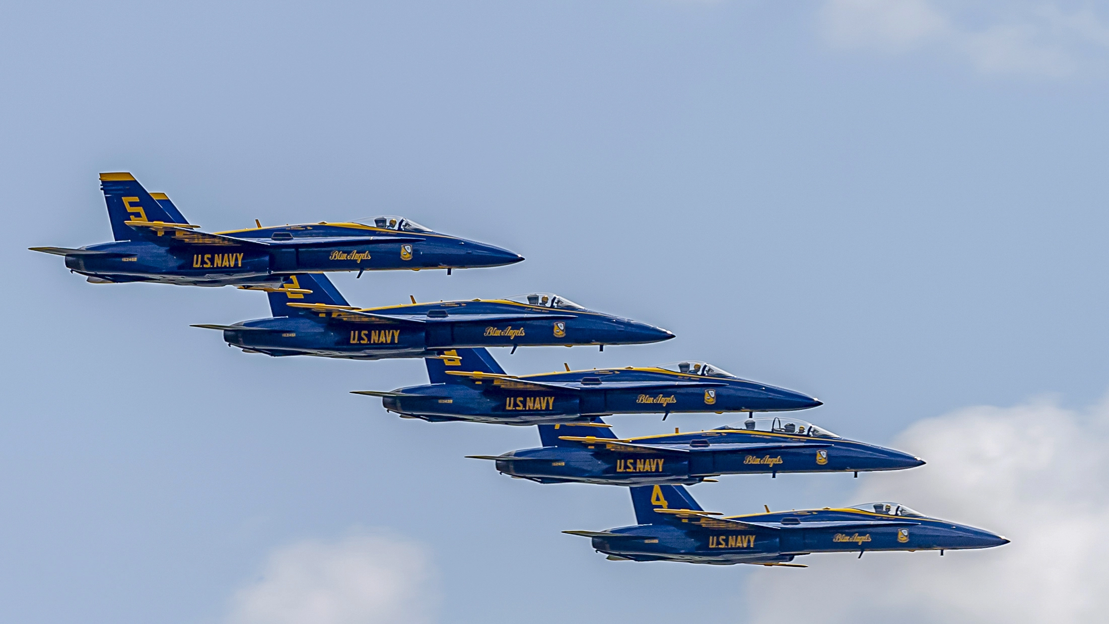Five U.S. Navy Blue Angels jets in a tight diamond formation flying against a blue sky with clouds.