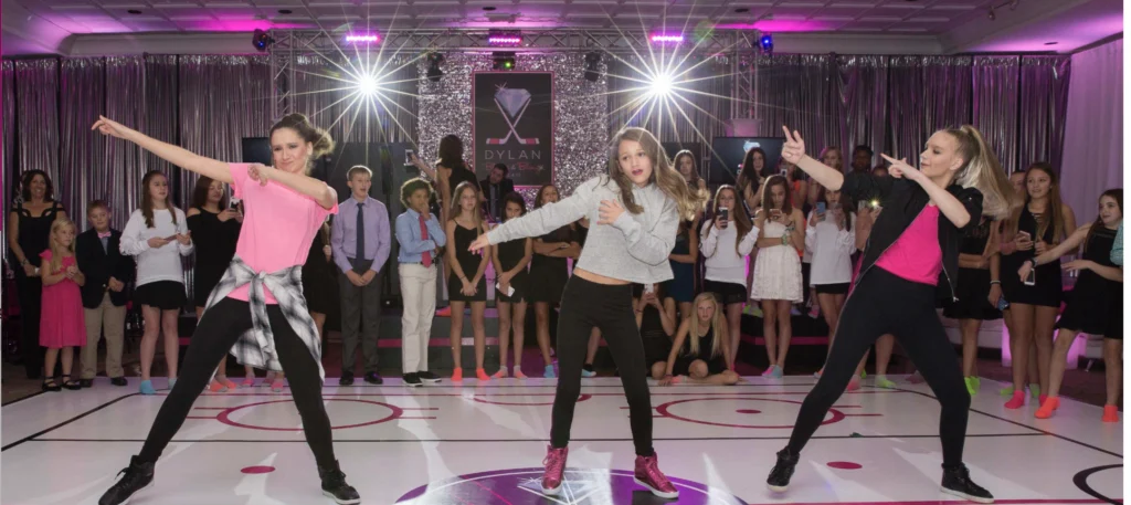 Three young women dance energetically on a stage with an audience watching. The setting has colorful lights and decorations.