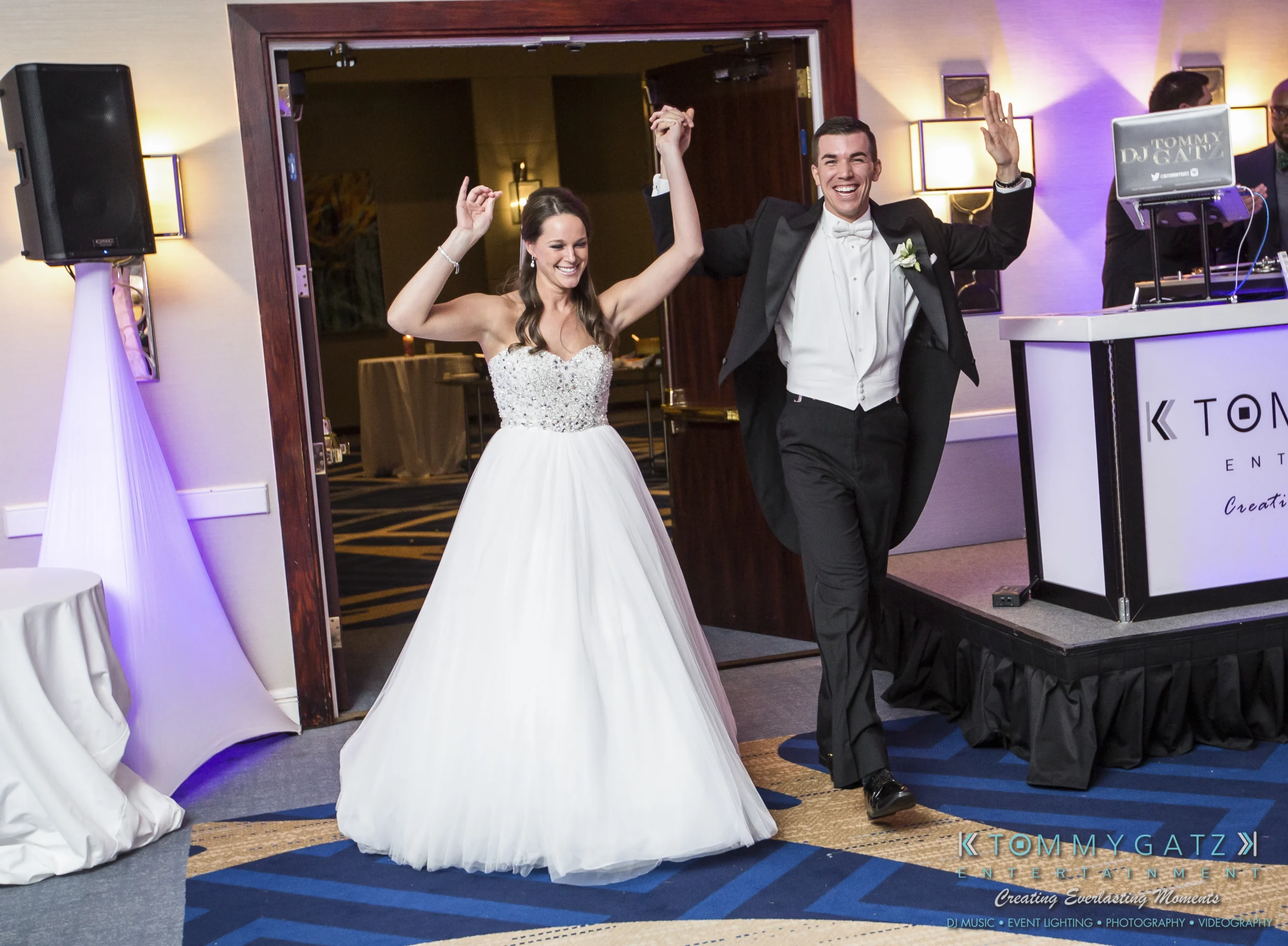 A bride and groom joyfully enter a reception hall, holding hands with arms raised. The groom wears a tuxedo, and the bride is in a white gown. A DJ booth is visible on the right.