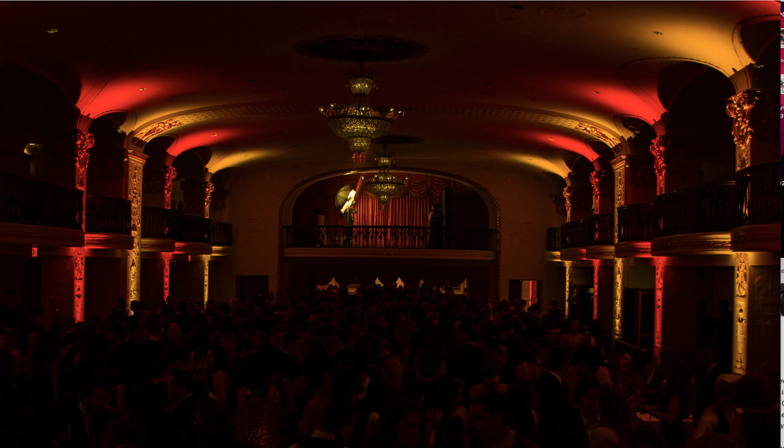 A dimly lit ballroom with red uplighting, chandeliers, and a crowd of people.