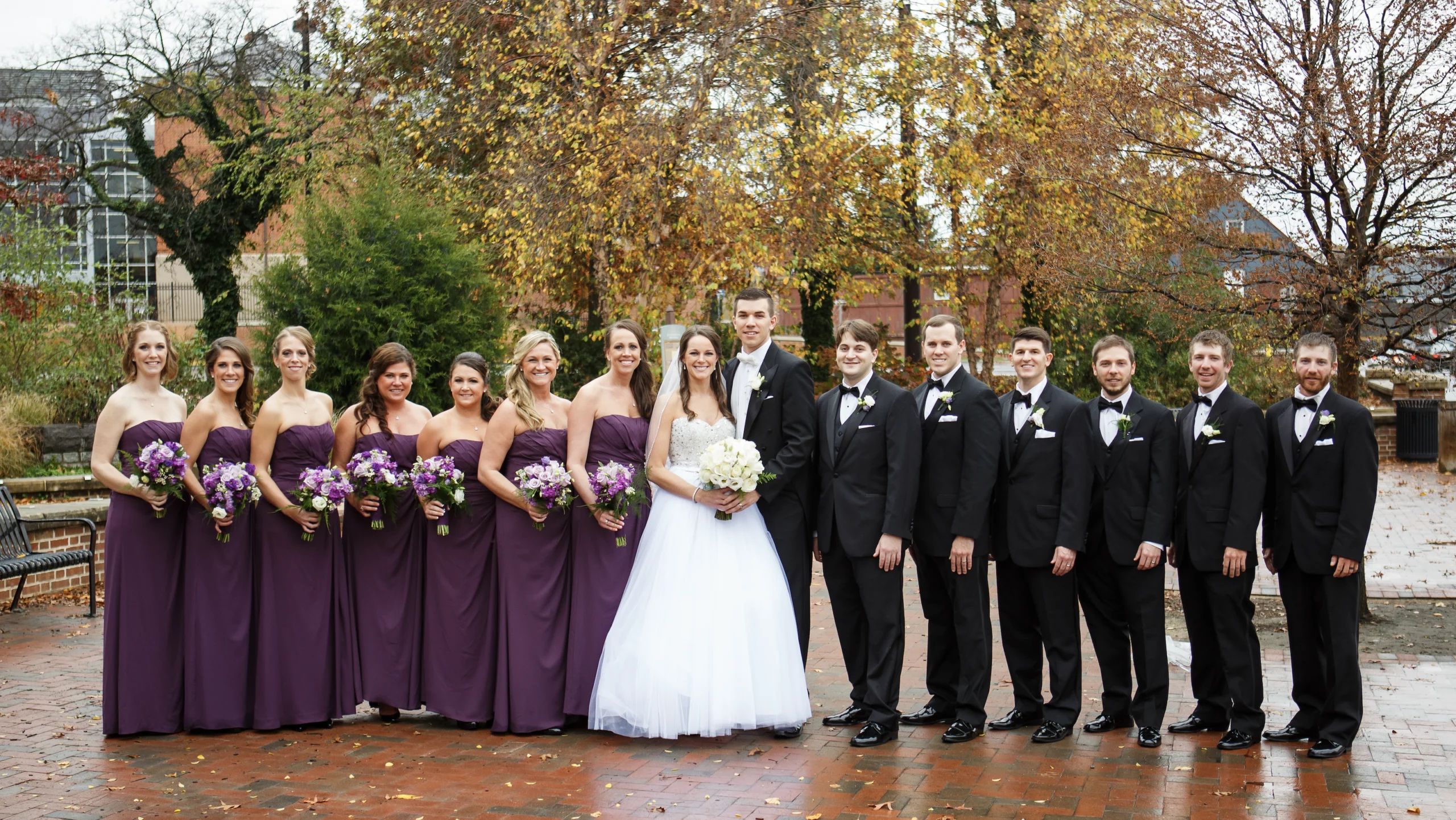 A bride and groom stand with eight bridesmaids in purple dresses and six groomsmen in black suits, posing outdoors on a wet brick path with trees in the background.