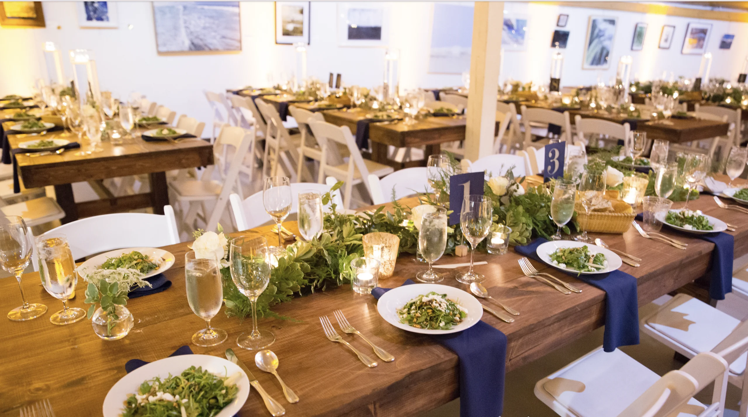 A wedding reception table set with plates of salad, glasses, cutlery, and candles on a wooden table with navy napkins and a greenery centerpiece. White chairs surround the table.