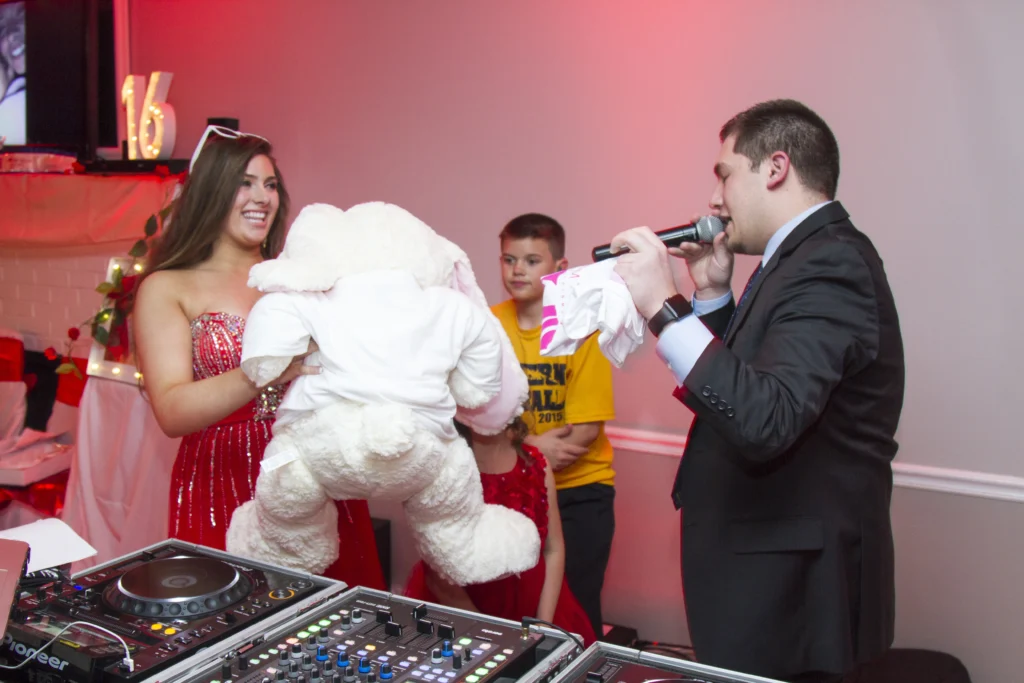 A man in a suit speaks into a microphone while a woman in a red dress holds a large teddy bear. A young boy in a yellow shirt stands beside them. Audio equipment is in the foreground.