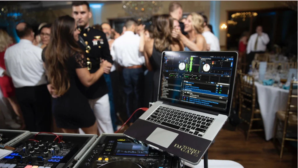 7PhotographyWeddings-Photo-_7-DJ-MusicOur-Complete- - TOMMY GATZ ENTERTAINMENT DJ setup with laptop displaying music software in the foreground. A crowd of people is dancing and socializing in the background.