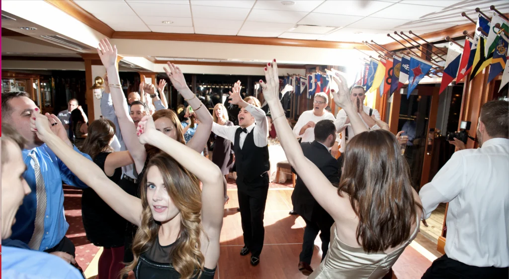 A group of people dancing energetically at a party, with their hands raised. The room is decorated with flags, and everyone appears to be enjoying the lively atmosphere.