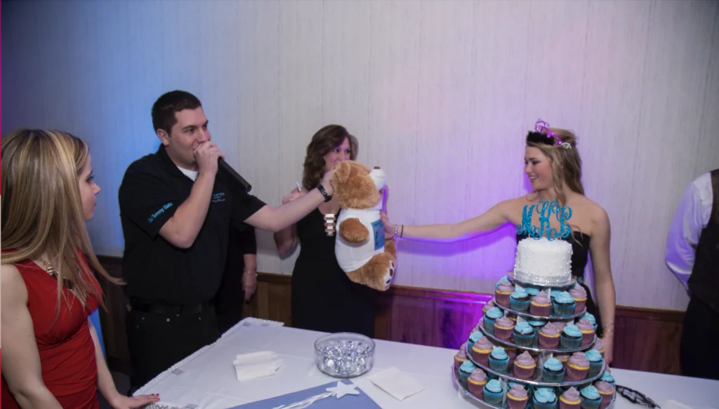 A woman in a tiara receives a teddy bear while a man speaks into a microphone at a celebration with a cake tower.