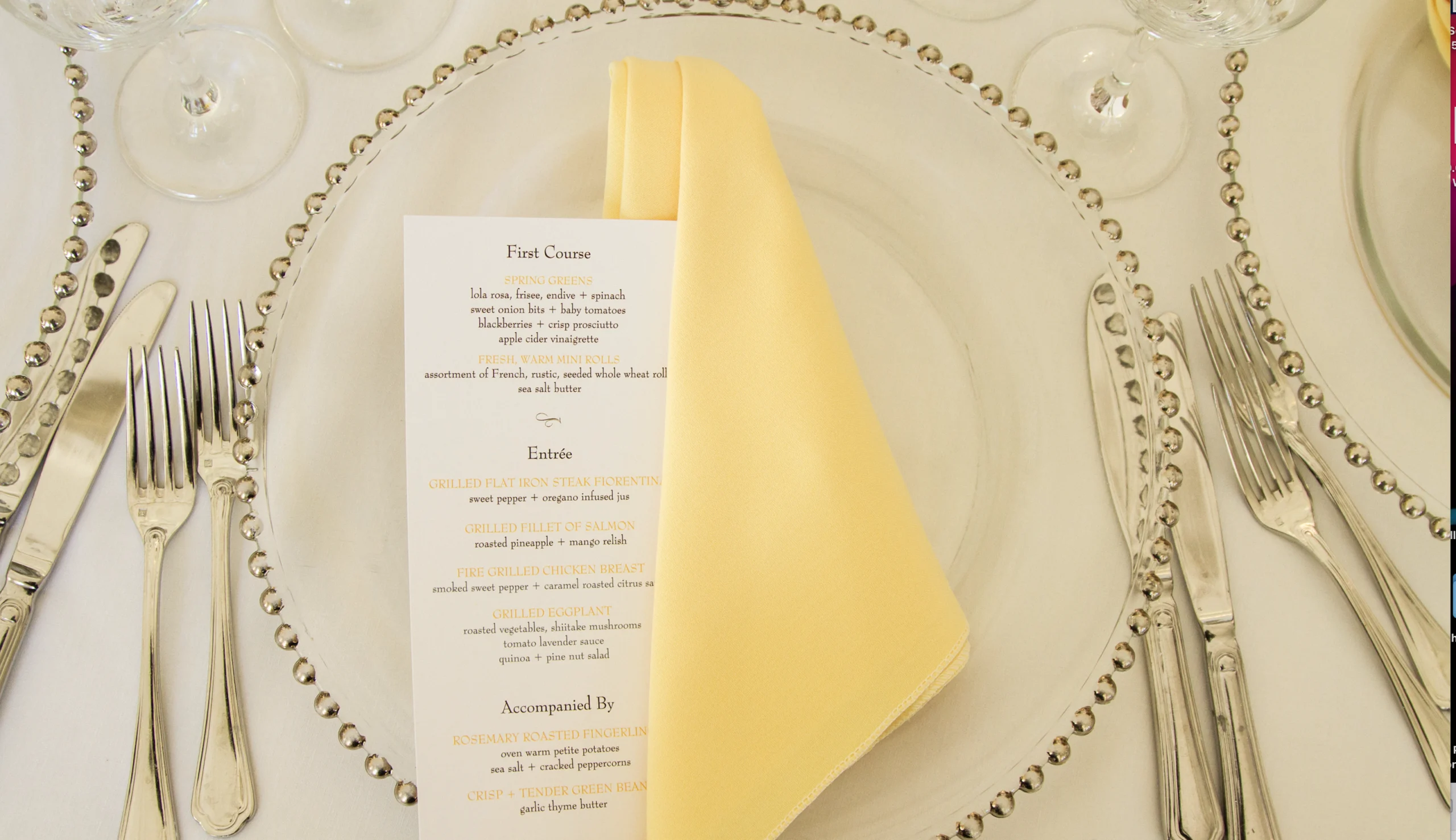 A dining place setting with a clear charger, yellow napkin, and a printed menu. Silverware surrounds the plate, and two wine glasses are positioned at the top.