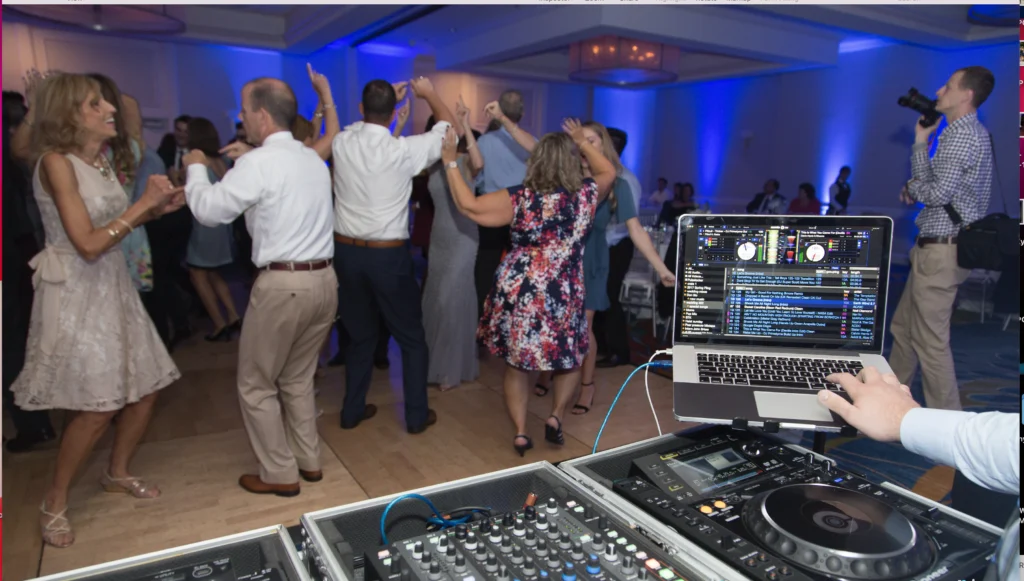 People dancing at an event with blue lighting, viewed from behind a DJ booth with a laptop and audio equipment in the foreground. A photographer is capturing the scene.