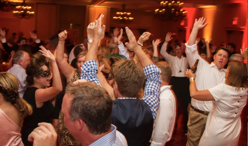 A lively crowd dances with raised hands at a party, some wearing masks, in a warmly lit room with chandeliers.