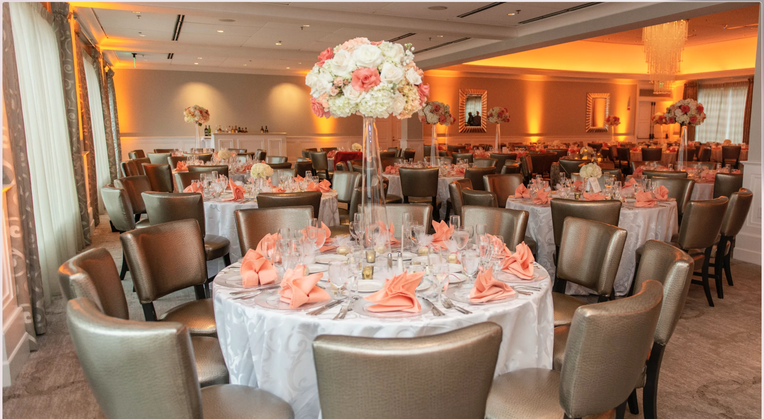 Elegant banquet hall with several round tables set for an event. Tables feature white tablecloths, peach napkins, glassware, and tall floral centerpieces. Soft lighting creates a cozy atmosphere.