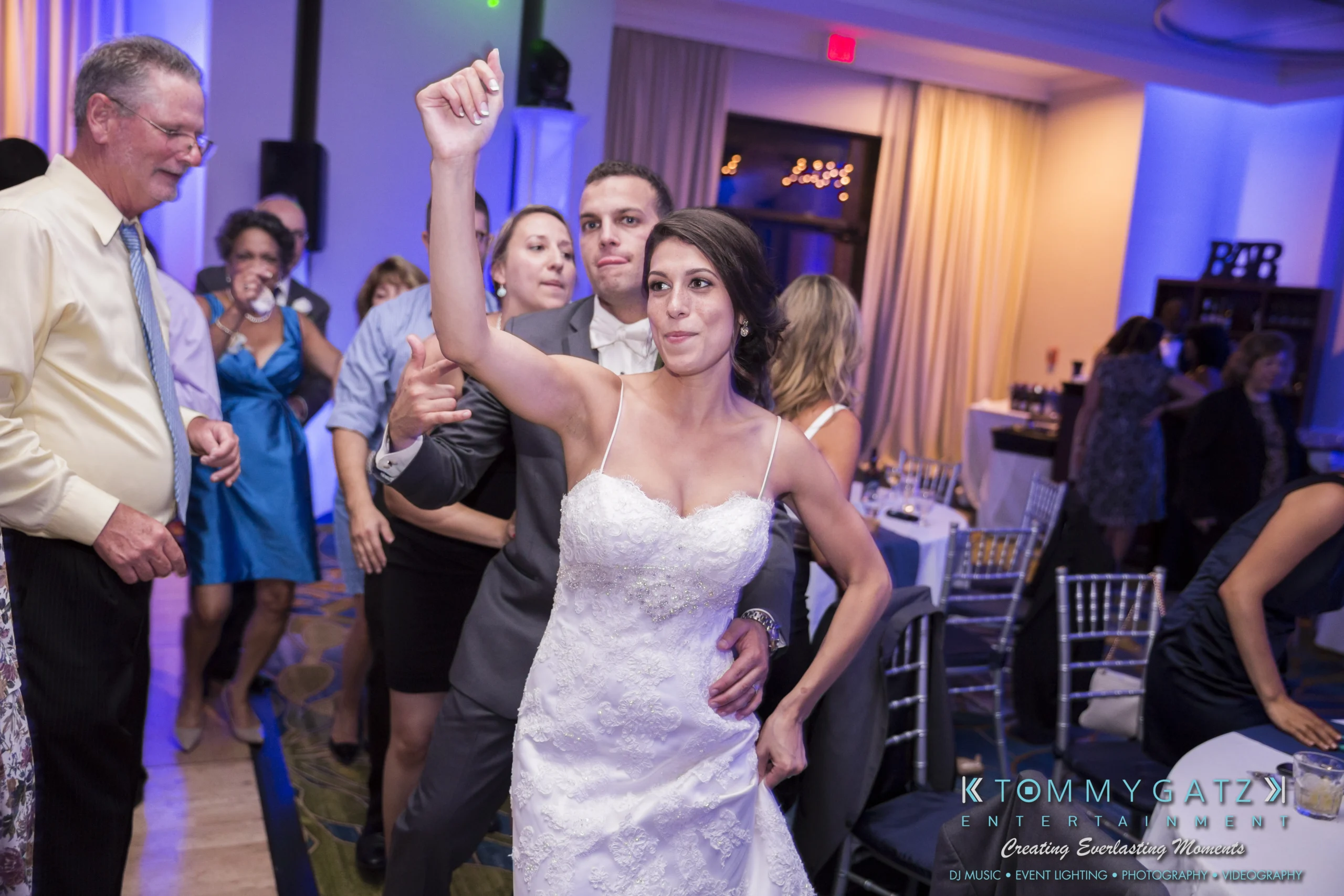 A bride in a white dress dances energetically at a reception, surrounded by smiling guests.