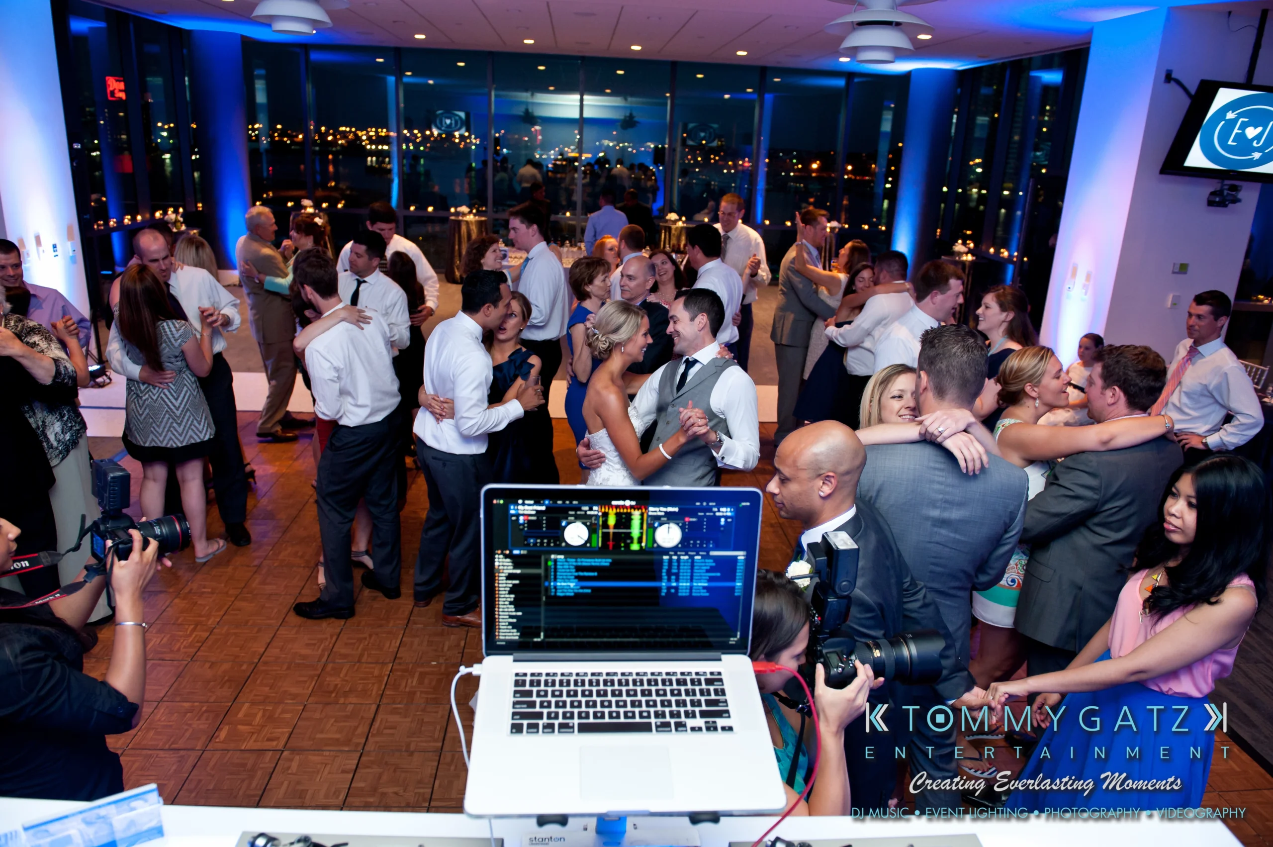People dancing and socializing at a lively event, with a DJ booth in the foreground and city lights visible through large windows in the background.