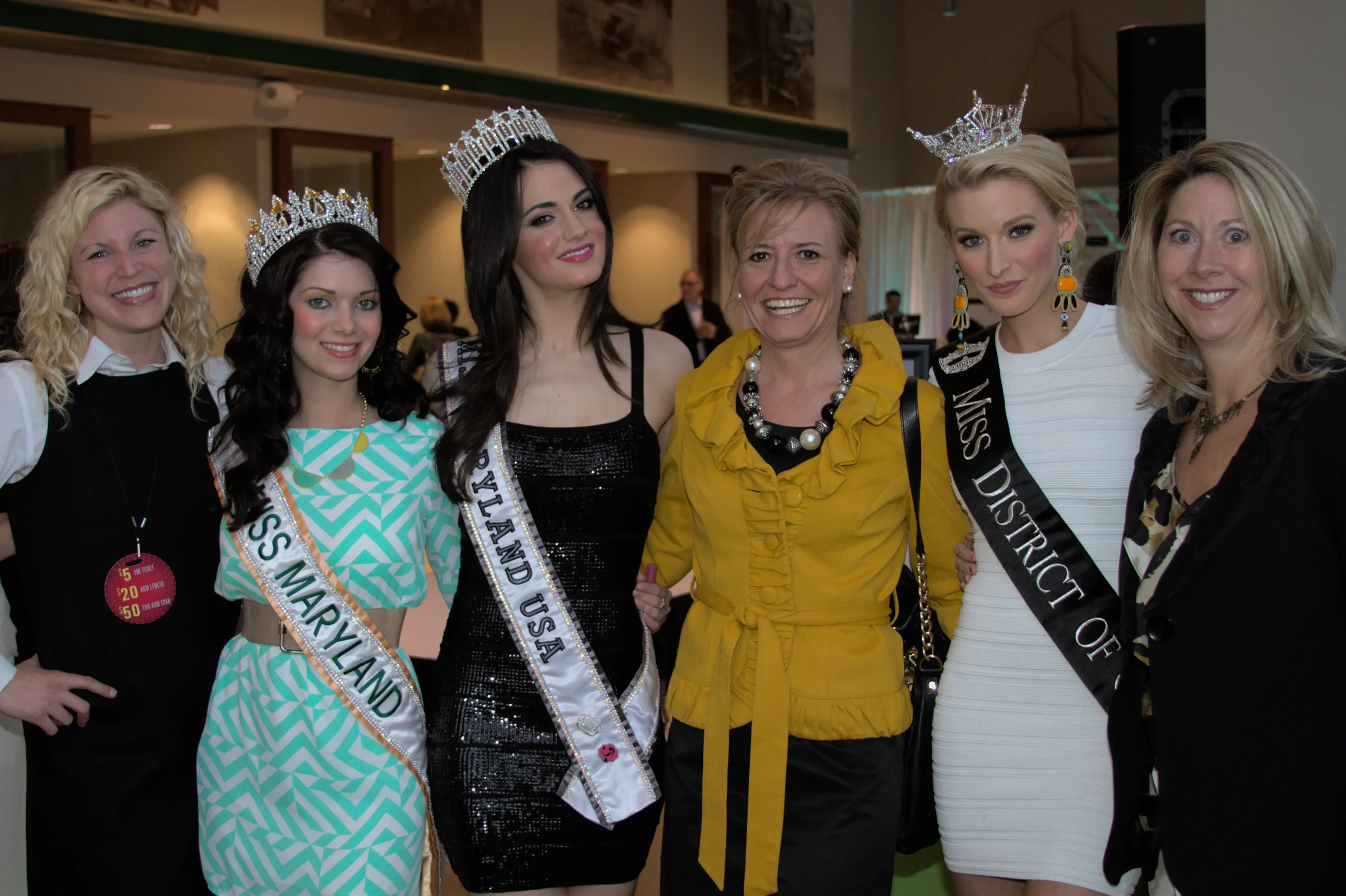 A group of six women smiling, including two wearing "Miss Maryland" sashes and one wearing "Miss District of Columbia" sash. Two are in crowns. They are indoors at an event.