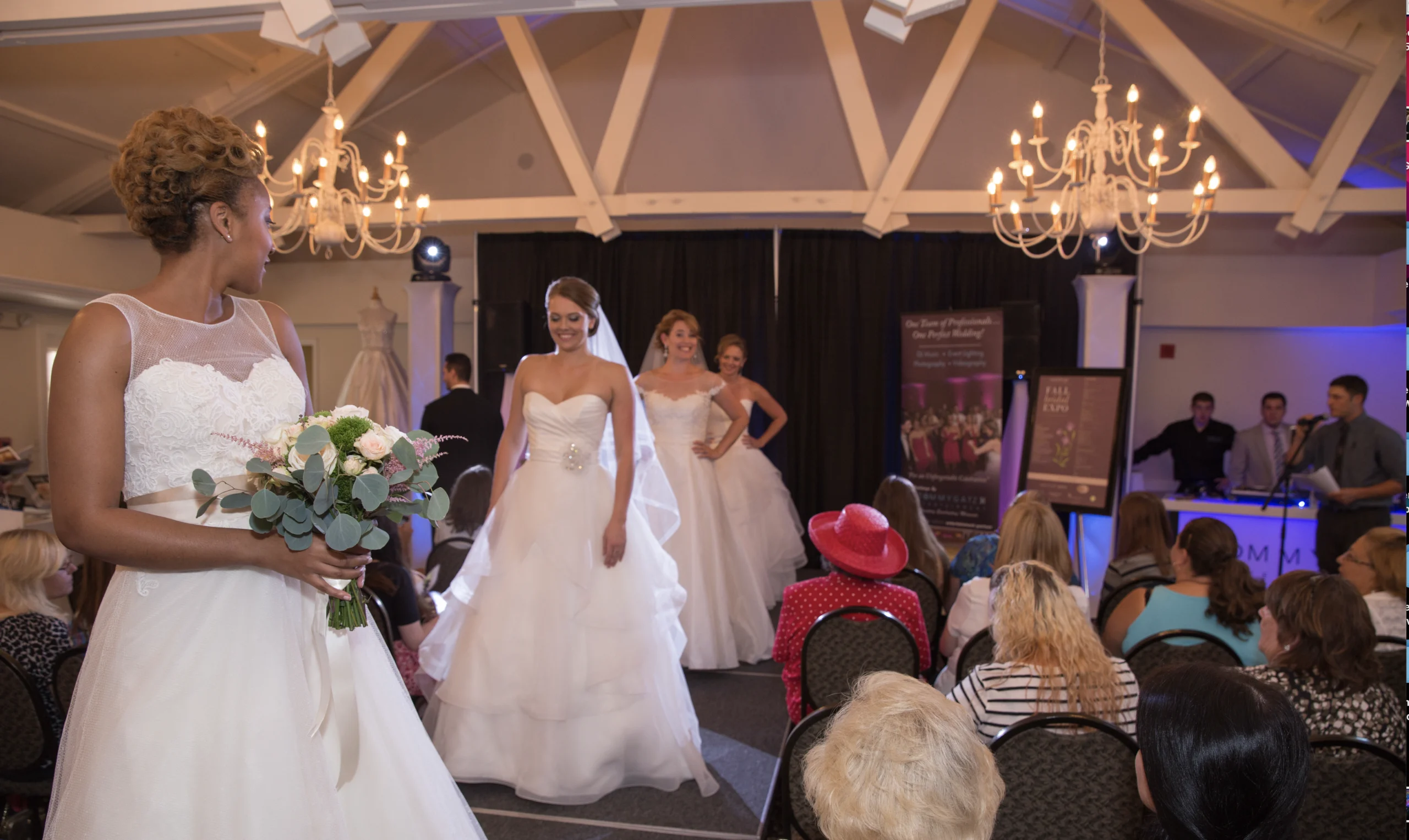 Bridesmaid holds bouquet while three models in wedding dresses walk on a runway. Audience seated, chandeliers hanging above.