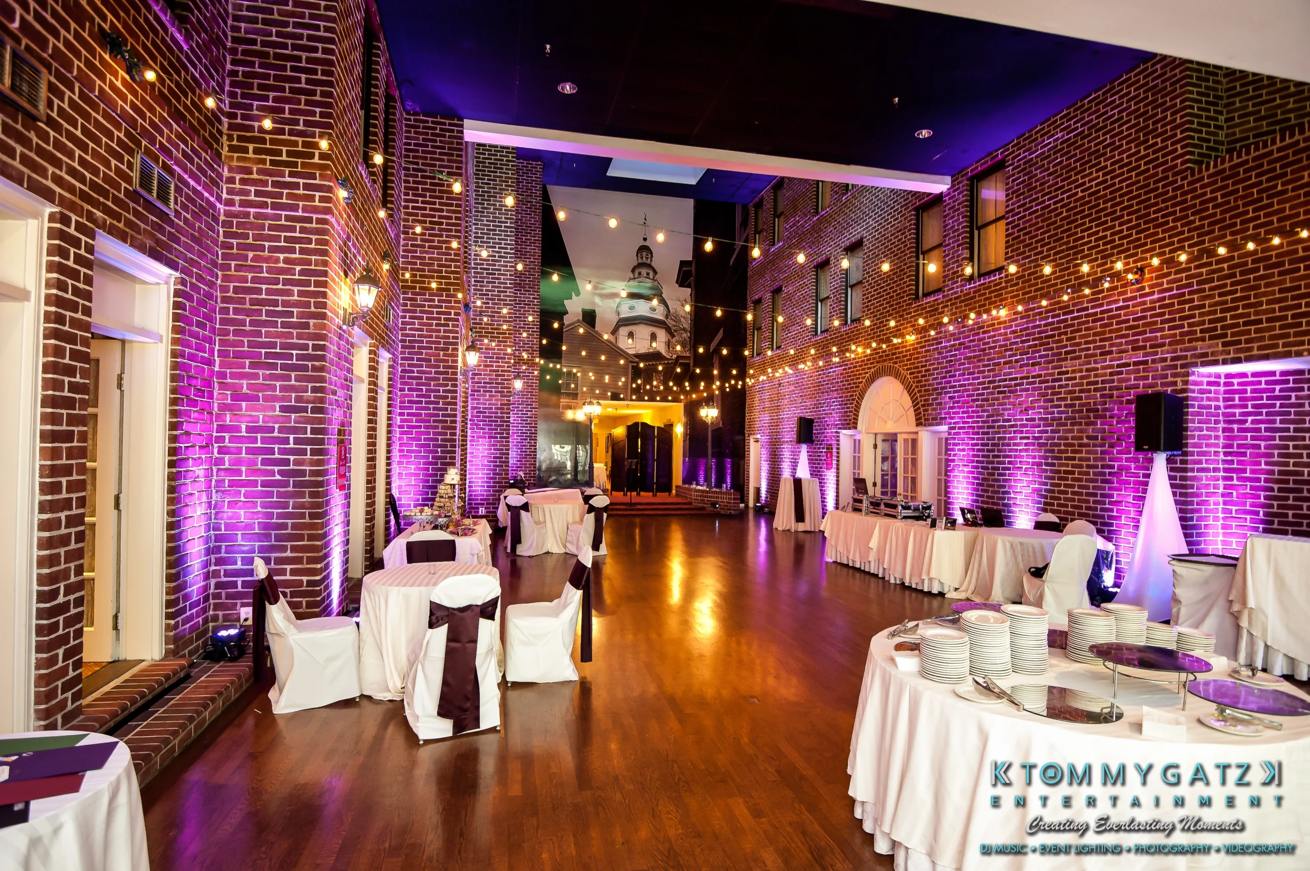 Spacious event hall with brick walls, string lights, and purple uplighting. Tables are set with white and purple linens. A logo is visible in the lower right corner.
