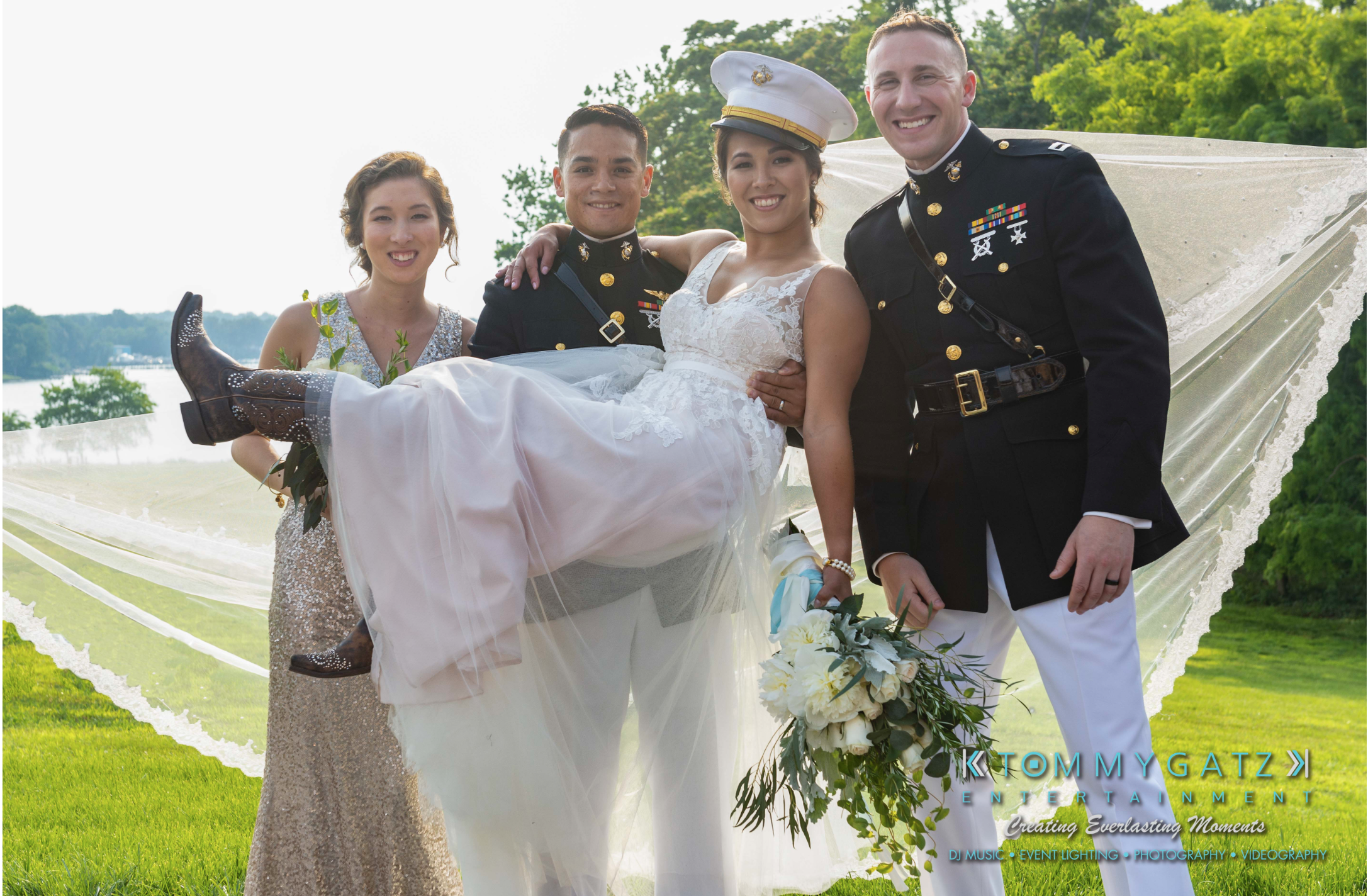 A bride in a cowboy hat and boots is playfully lifted by two men in military uniforms, standing beside a smiling bridesmaid. They are outdoors with a lake and greenery in the background.
